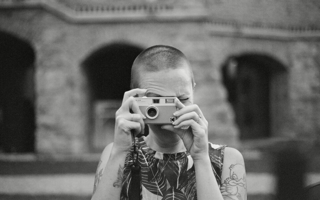 black and white image of woman with shaved head in a floral top taking a photo with a film camera